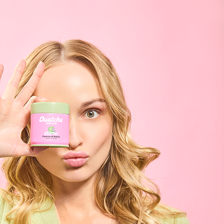 Woman holding a pink Ouatcha Matcha tin with a green lid against a pink background.