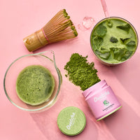 Flat lay of prepared matcha, bamboo whisk, and a Daily Ouatcha Matcha ceremonial-grade first harvest matcha tin on a pink background.