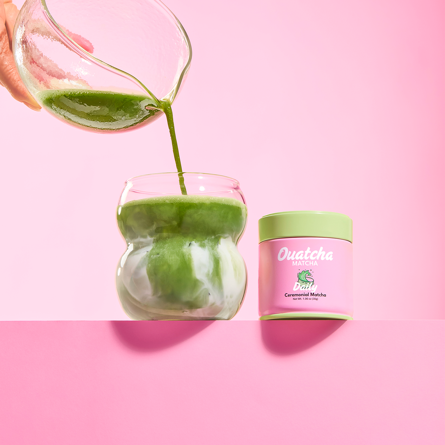 Green ceremonial first harverst matcha being poured into a glass next to a 'Daily' Ouatcha Matcha tin on a pink background.