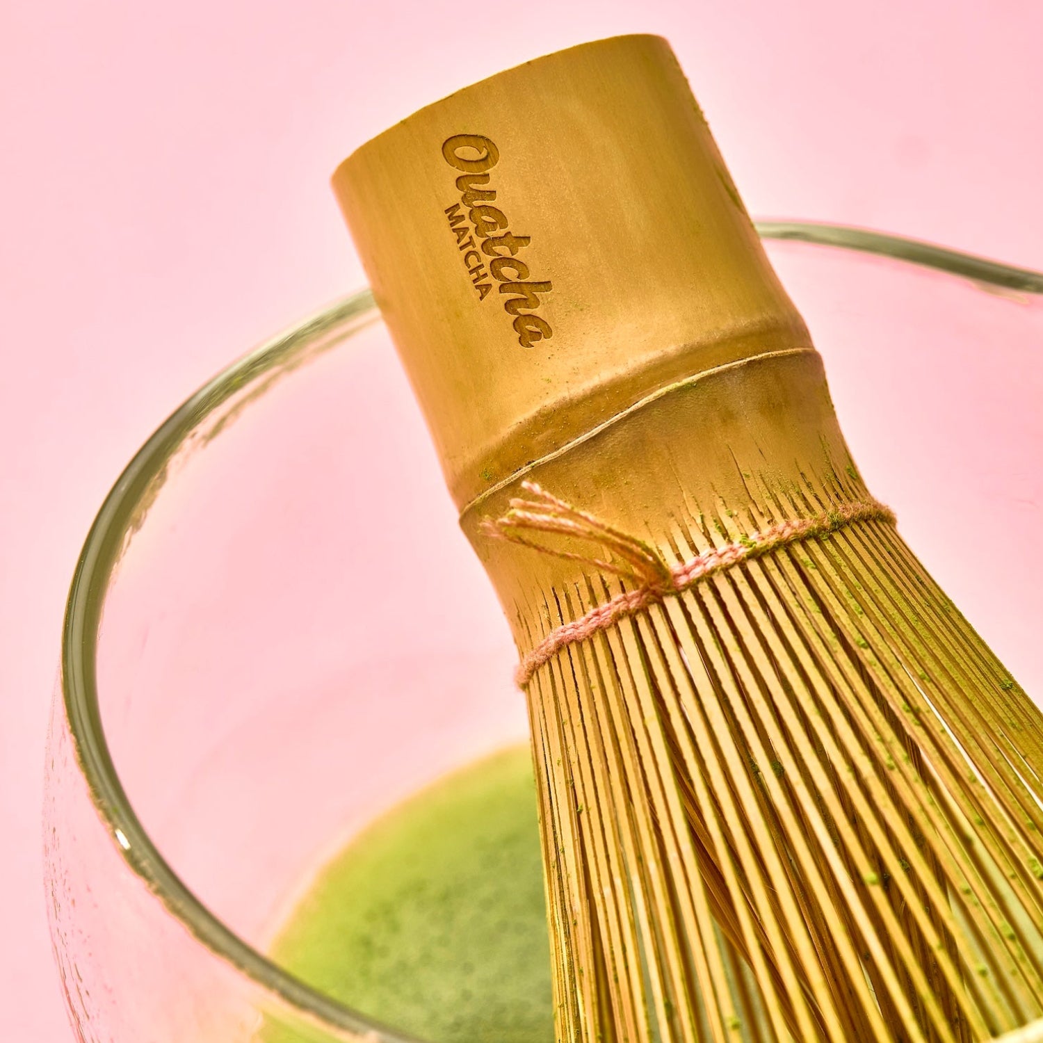 Close-up of an Ouatcha Matcha bamboo whisk resting in a glass of prepared matcha on a pink background
